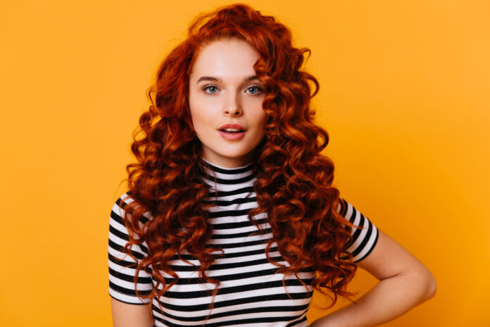Close-up portrait of blue-eyed girl with long eyelashes and voluminous red curly hair against orange background.